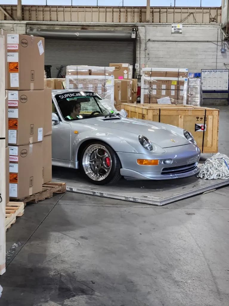 A silver Porsche 911 being prepared for shipment inside a warehouse, surrounded by shipping crates and boxes.