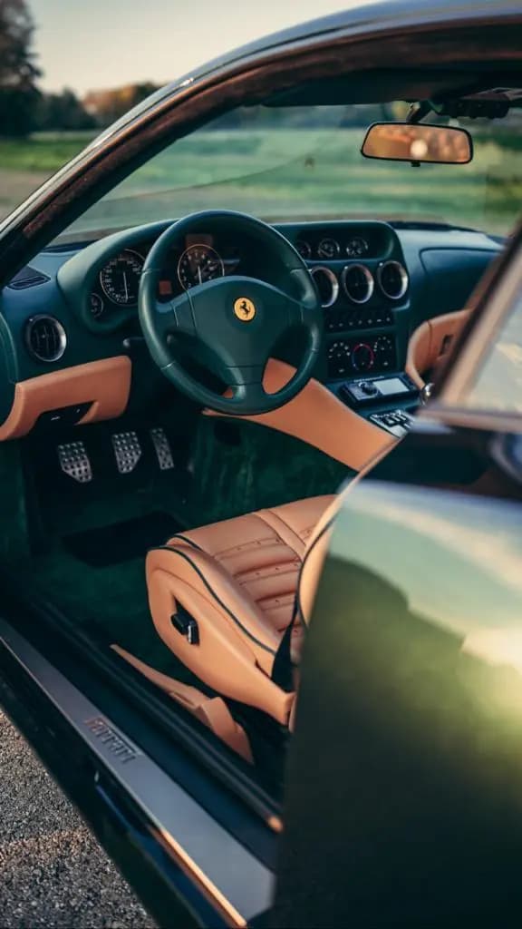 Interior view of a luxury sports car with tan leather seats, a green steering wheel featuring the Ferrari logo, and the driver's side door open.